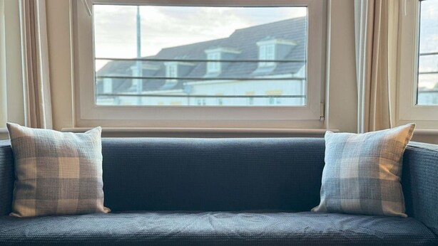 A dark blue sofa with two plaid pillows, positioned in front of a window with a view of buildings. - Kingfisher Apartment (Fife)