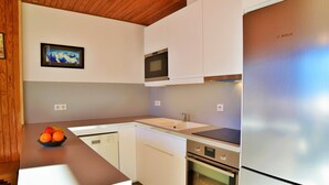 A modern kitchen with white cabinetry, a stainless steel refrigerator, a microwave, and an oven. There is a fruit bowl on the counter and a framed picture on the wall.