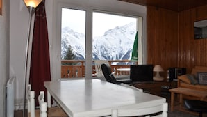 A room with a large window overlooking snow-capped mountains, a table, chairs, a TV, and a couch.