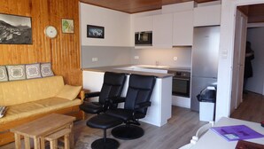 A compact living space with a sofa, two black chairs, a small wooden table, a kitchen area with a sink, stove, and refrigerator, and a clock on the wall.