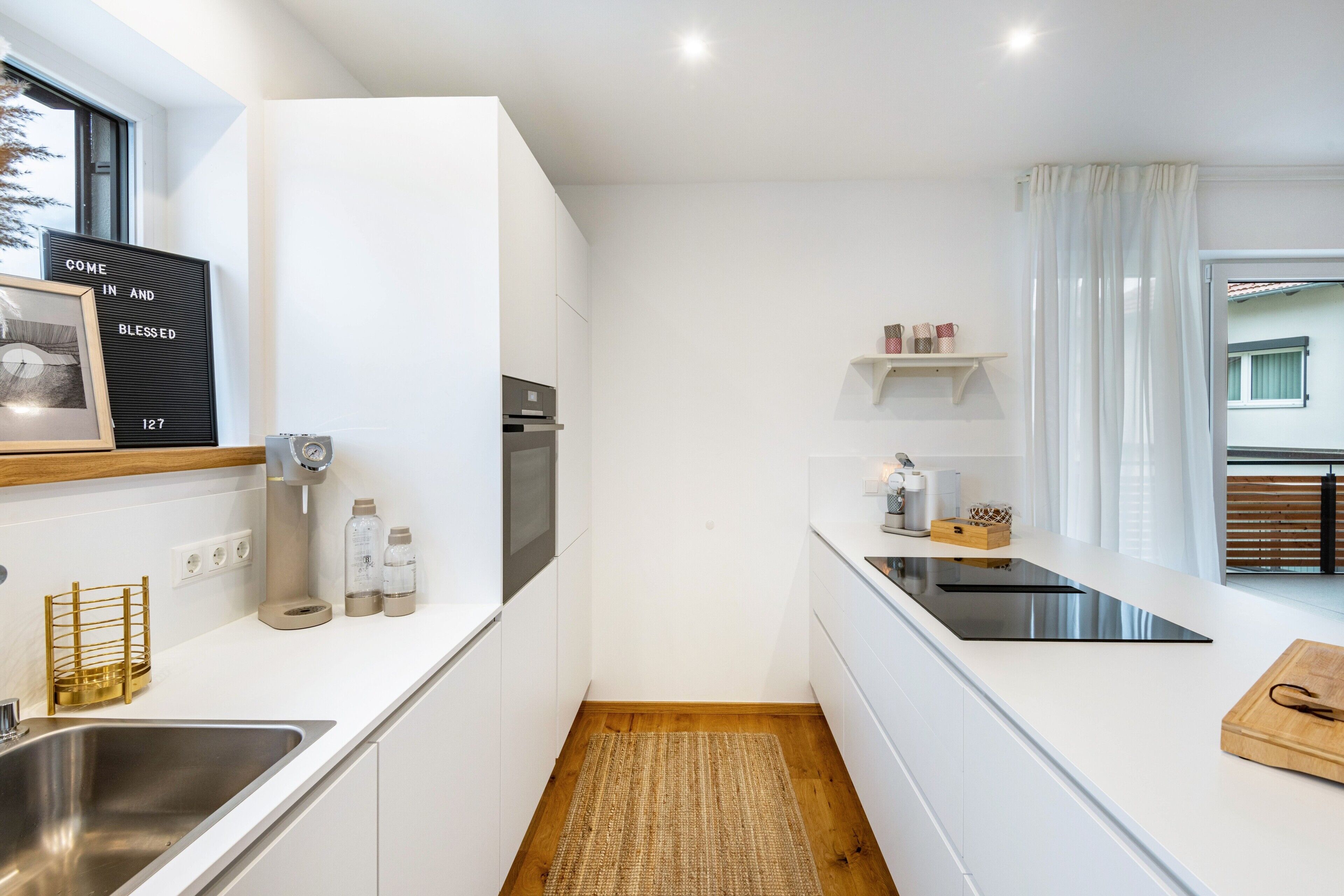 Una cocina moderna con muebles blancos, un lavamanos de acero inoxidable, horno integrado y suelo de madera.