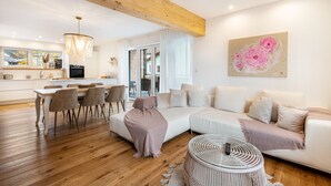 A modern living room with a sofa, dining table, and chairs. There is a kitchen in the background with a large window. - Holiday flat ‘Bei Thérèse’ with balcony and Wi-Fi (Miltach)