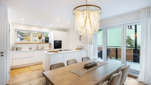 A modern kitchen with white cabinets, a dining area with a wooden table, and a large window overlooking a balcony. - Holiday flat ‘Bei Thérèse’ with balcony and Wi-Fi (Miltach)