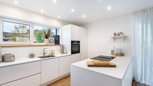 A modern kitchen with white cabinetry, a built-in oven, a microwave, and a large window with a view of trees and buildings. - Holiday flat ‘Bei Thérèse’ with balcony and Wi-Fi (Miltach)