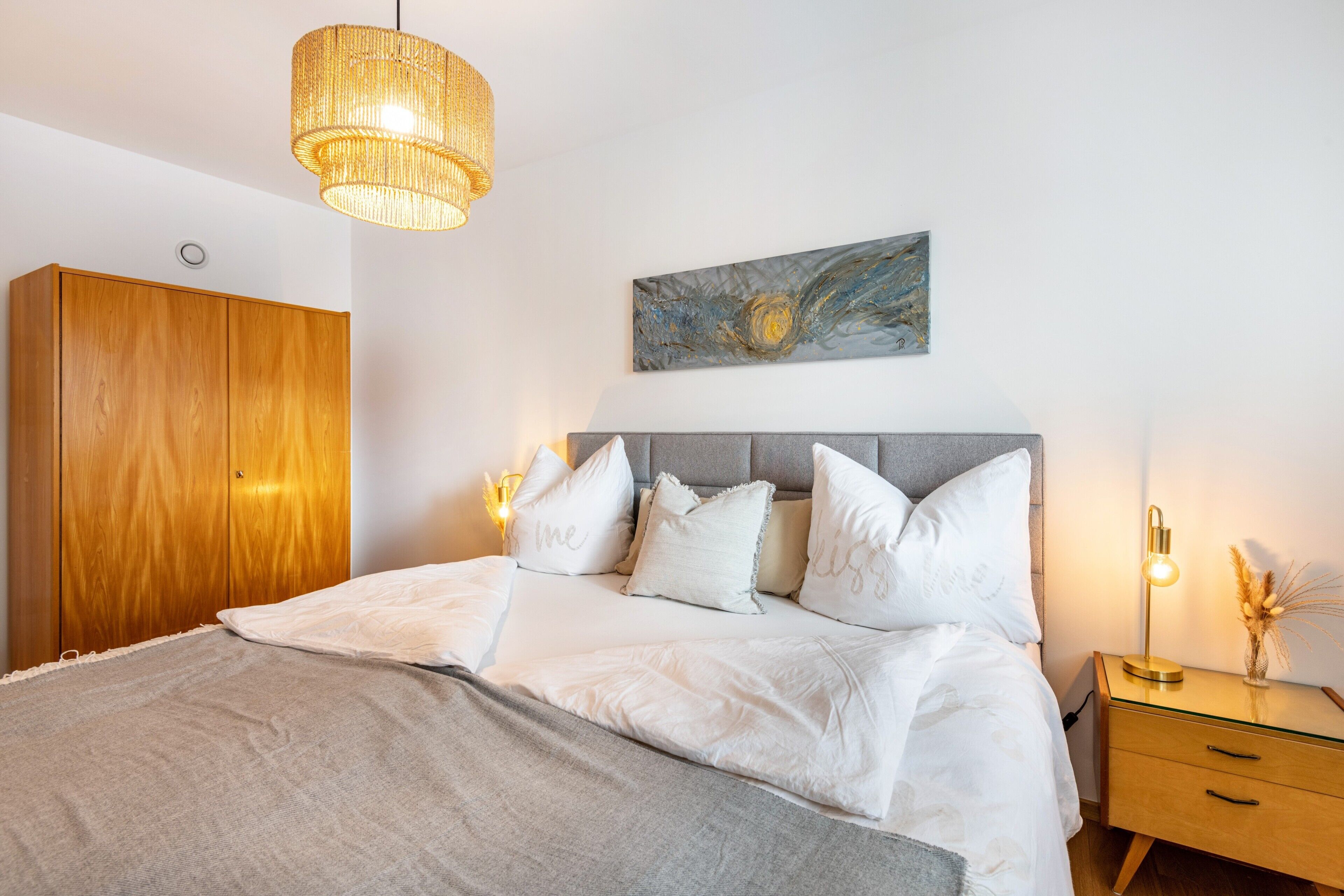 Une chambre à coucher comprenant un lit, des lampes de chevet, une armoire en bois, une tête de lit et une œuvre d’art accrochée au mur.