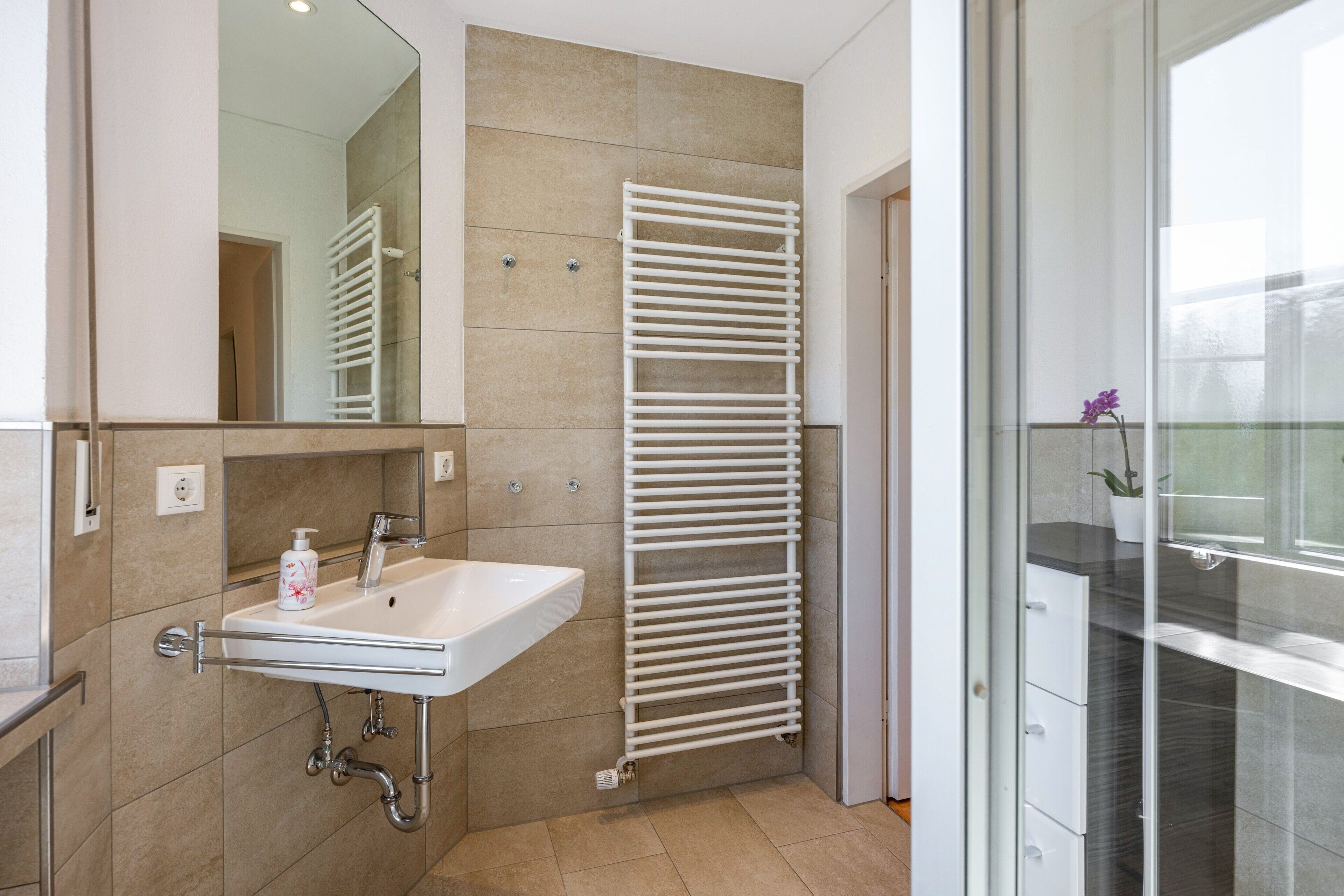 A modern bathroom with a white sink, a mirror, a towel rack, and a glass door leading to a balcony with a potted plant.
