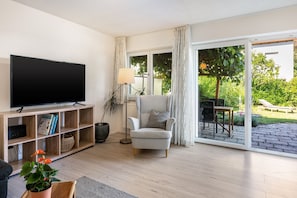 A living room with a flat-screen TV on a wooden stand, a grey armchair, a floor lamp, and a large sliding glass door leading to an outdoor area with a table and chairs.