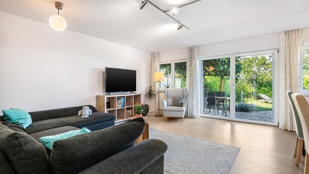 Ein modernes Wohnzimmer mit einem Flachbildfernseher, grauen Sofas, einem Holzregal und Blick auf einen Garten durch große Schiebeglastüren.