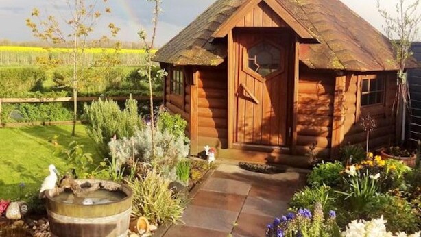 Une cabane en bois avec un toit de chaume, entourée d’un jardin où poussent différentes plantes et près d’un petit étang.