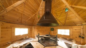 L’intérieur d’un chalet en bois, avec un poêle au centre, des bancs recouverts de fourrure et des fenêtres.