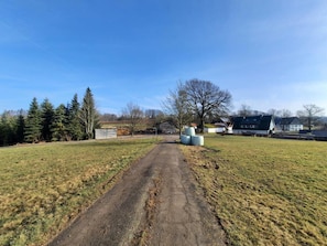 A rural road leading to a building with a green roof, surrounded by trees and open fields.