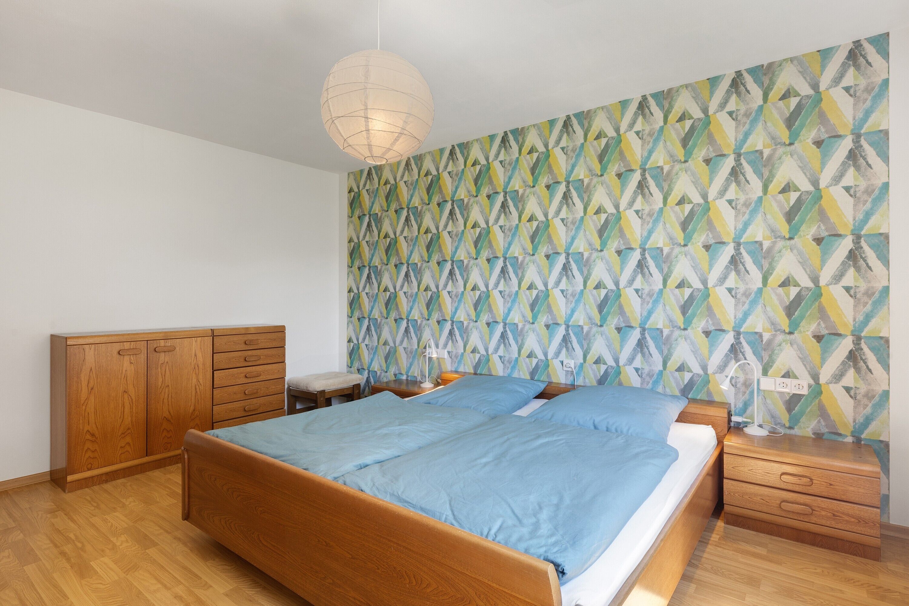 Quarto com cama, guarda-roupa de madeira, mesas de cabeceira e parede com padrão geométrico.