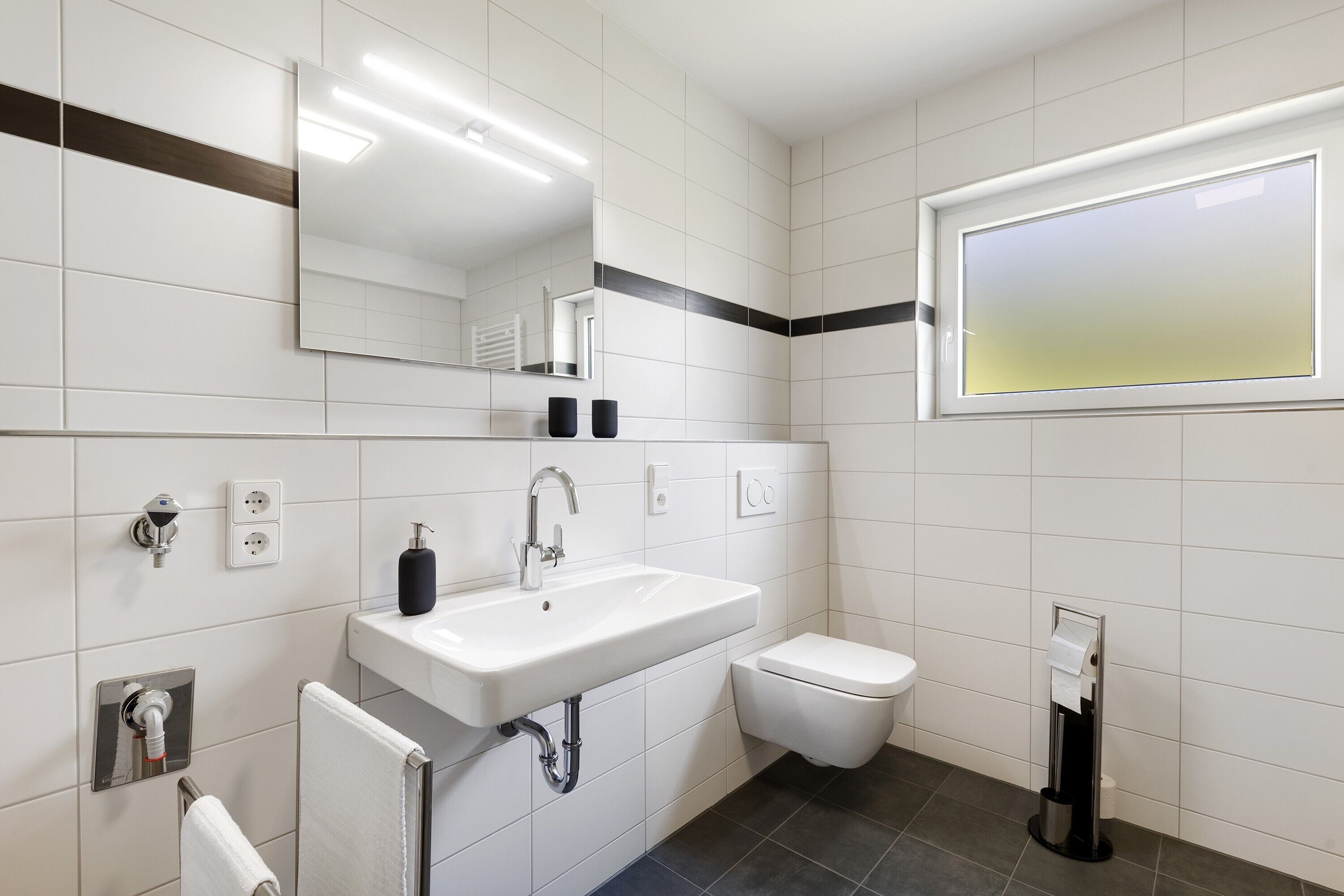 Un baño moderno con un lavamanos blanco, un inodoro, un espejo y una ventana.
