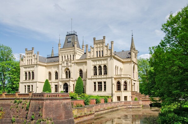 A large, historic building with multiple towers and arched windows, surrounded by greenery and a water feature.
