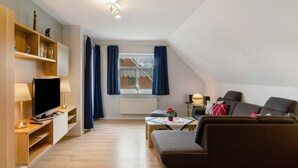 Una sala de estar moderna con un televisor de pantalla plana, estanterías de madera, un sofá, una mesa con un jarrón y una ventana con cortinas.