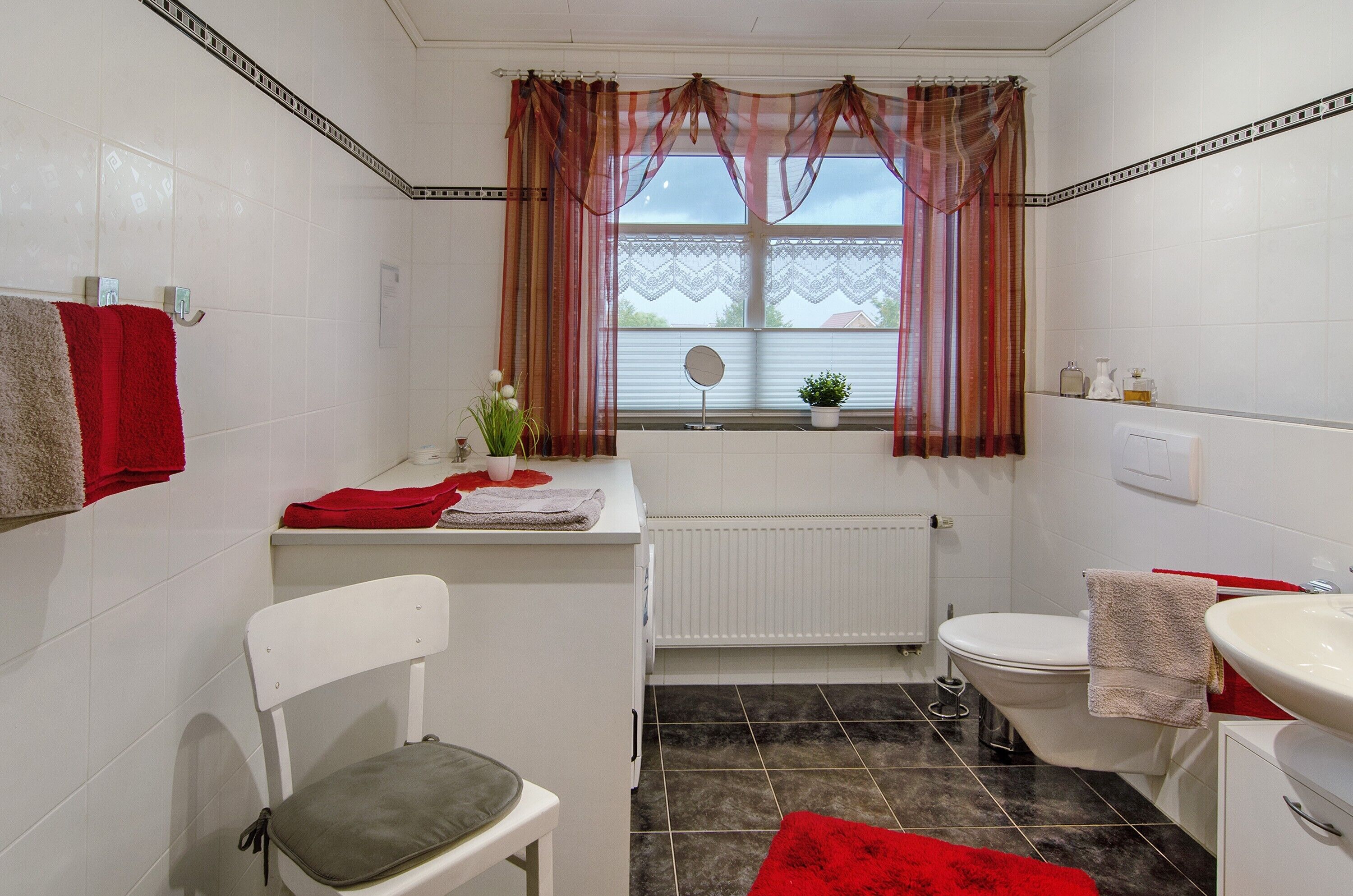 A bathroom with a white radiator, a toilet, a sink, a chair, and a window with curtains.