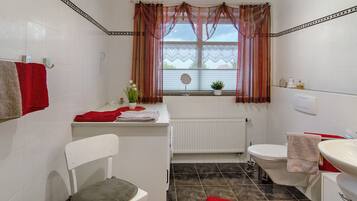 A bathroom with a white radiator, a toilet, a sink, a chair, and a window with curtains.