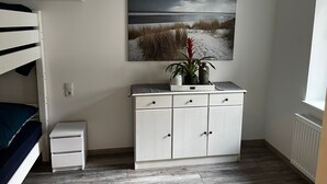 Une chambre avec un lit superposé, une commode blanche à tiroirs, une plante en pot et un tableau représentant un paysage de plage accroché au mur.
