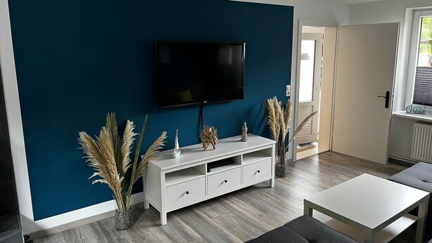Un salon moderne avec un mur d’accent bleu foncé, un téléviseur à écran plat, un meuble de rangement blanc, un canapé gris et une table basse blanche.