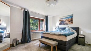 Une chambre à coucher comprenant un lit, un banc, une table de chevet, une fenêtre avec des rideaux, un radiateur et un tableau accroché au mur.