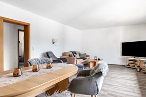 Uma sala de estar moderna com uma mesa de jantar de madeira, cadeiras estofadas cinzentas, um sofá e uma televisão de tela plana.