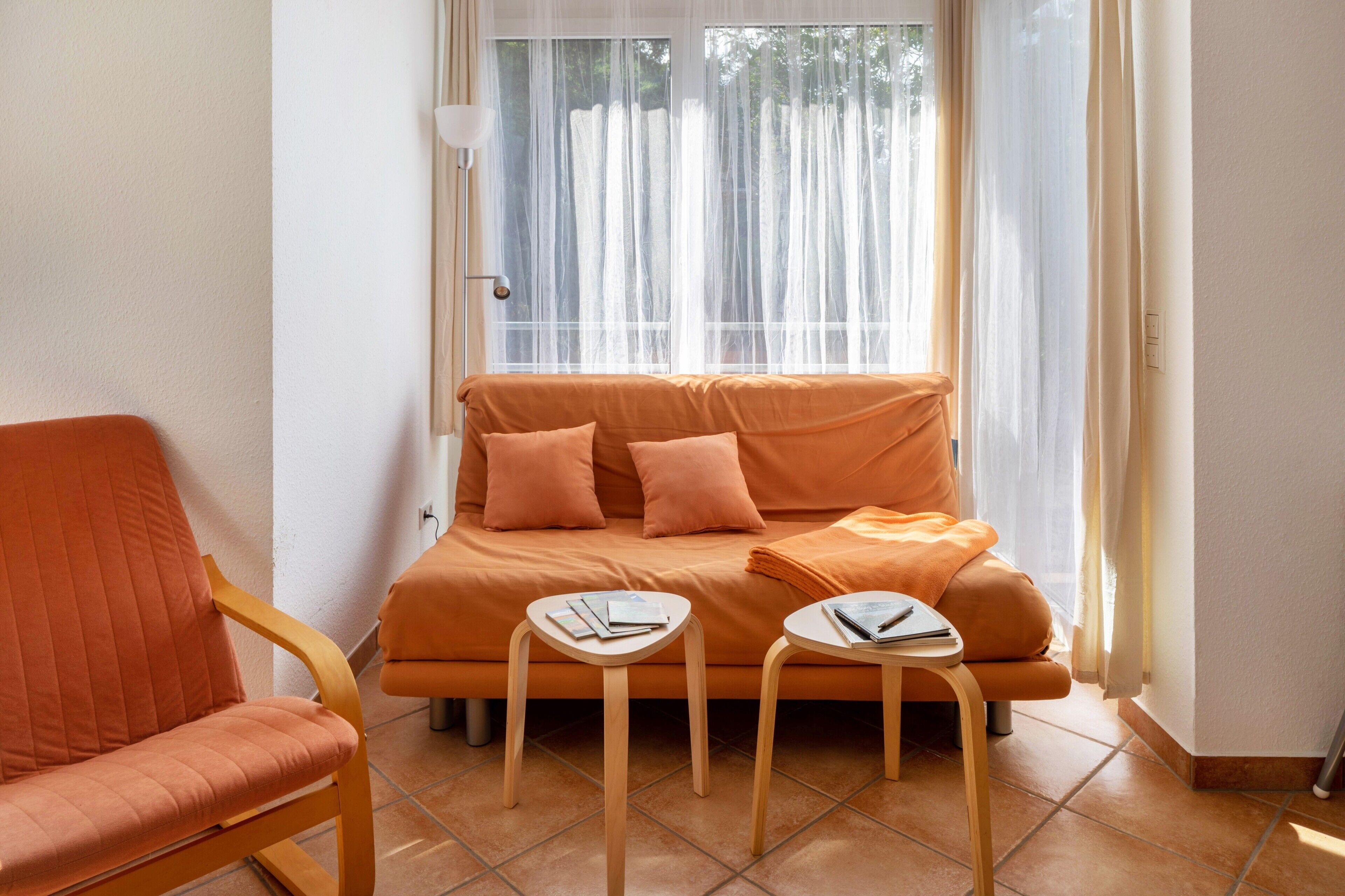 Huoneessa on sohva, kaksi pientä pöytää, tuoli ja ikkuna, jossa on ohuet verhot.