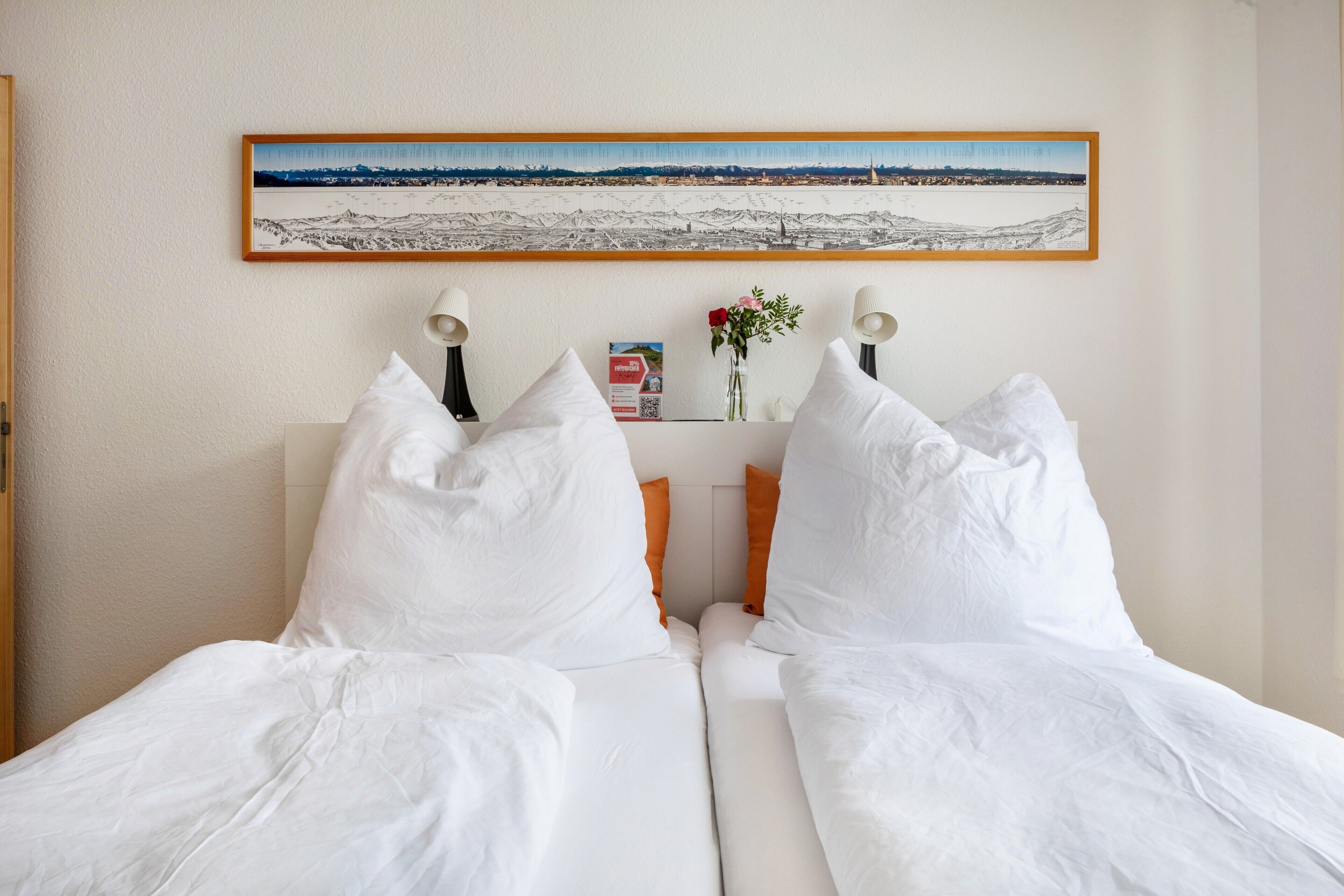 A bed with white bedding, two bedside lamps, a framed landscape painting, and a small vase with flowers.