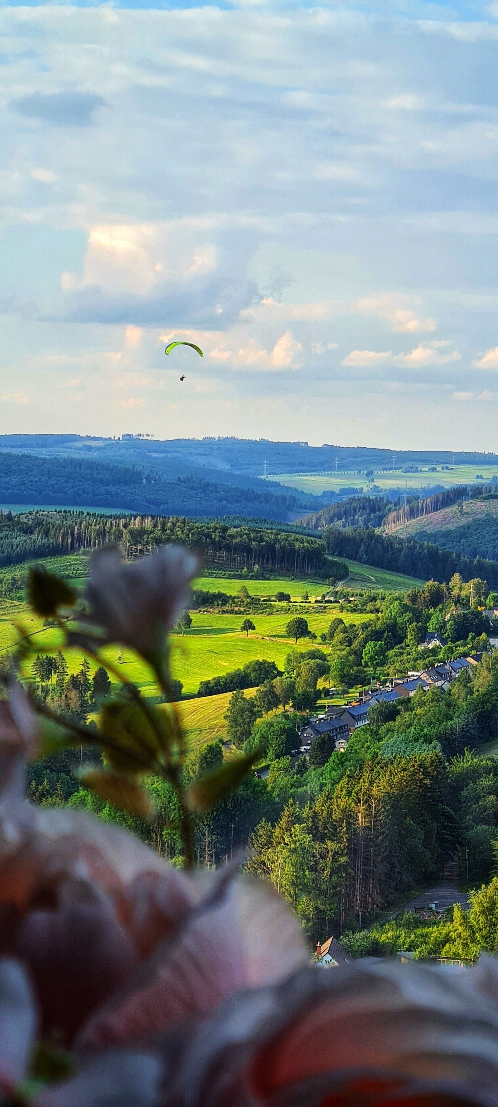 Een landschap met een groen veld, een huis en een persoon die paraglideert in de verte.