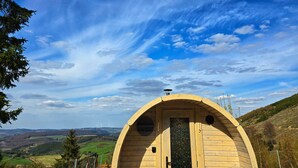 Una cabaña de madera con techo curvo, una puerta y una ventana, con un cielo despejado y un paisaje montañoso de fondo.