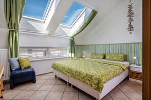 Un dormitorio con tragaluz, una cama con ropa de cama verde, una silla con cojines y una mesita de noche con una lámpara.