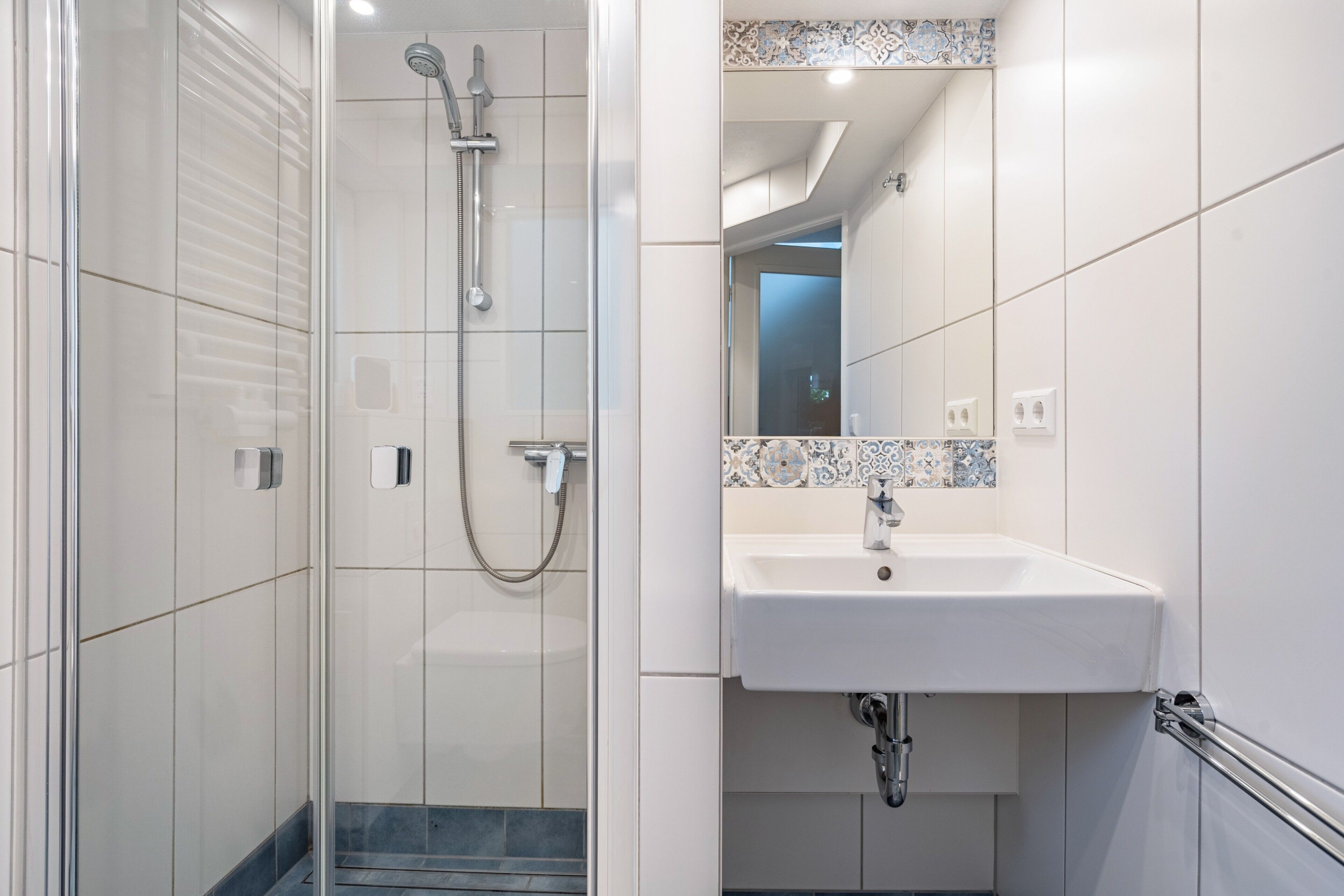 Ein modernes Badezimmer mit einer gläsernen Duschkabine, einem weißen Waschbecken mit gefliestem Rücksprung und einem Spiegel darüber.