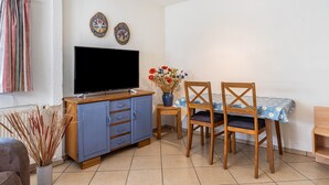 Ein Zimmer mit Fernseher, einem Esstisch mit Stühlen, einem kleinen Schrank und einer Vase mit Blumen.