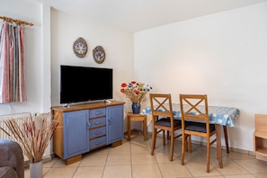 A room with a television, a dining table with chairs, a small cabinet, and a vase with flowers.