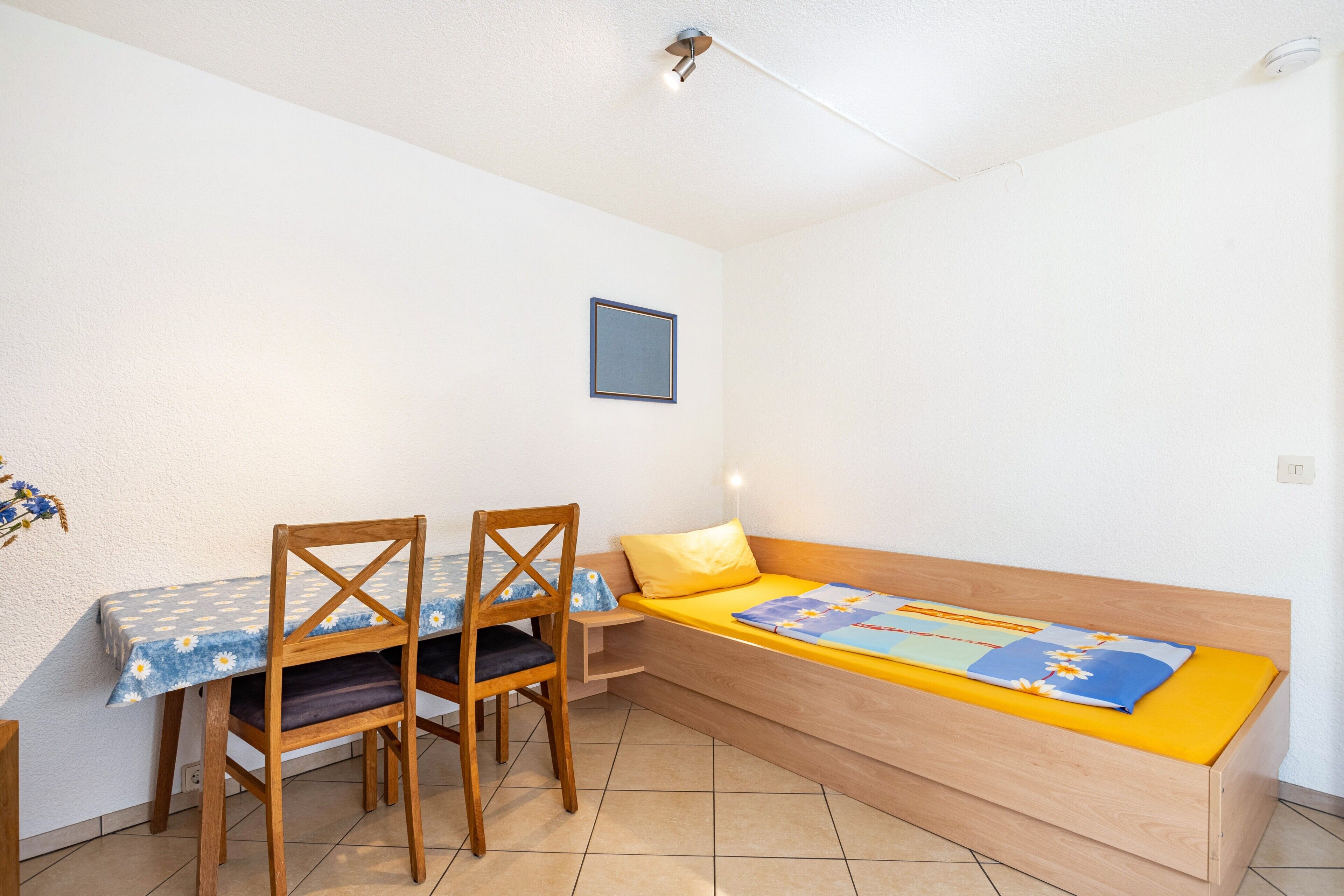 Une petite pièce avec un lit en bois, une table avec des chaises et un seul luminaire fixé au mur.