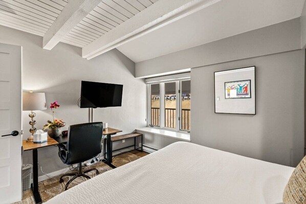 A hotel room with a bed, a desk with a chair, a television, a window with a view, and a framed picture on the wall.