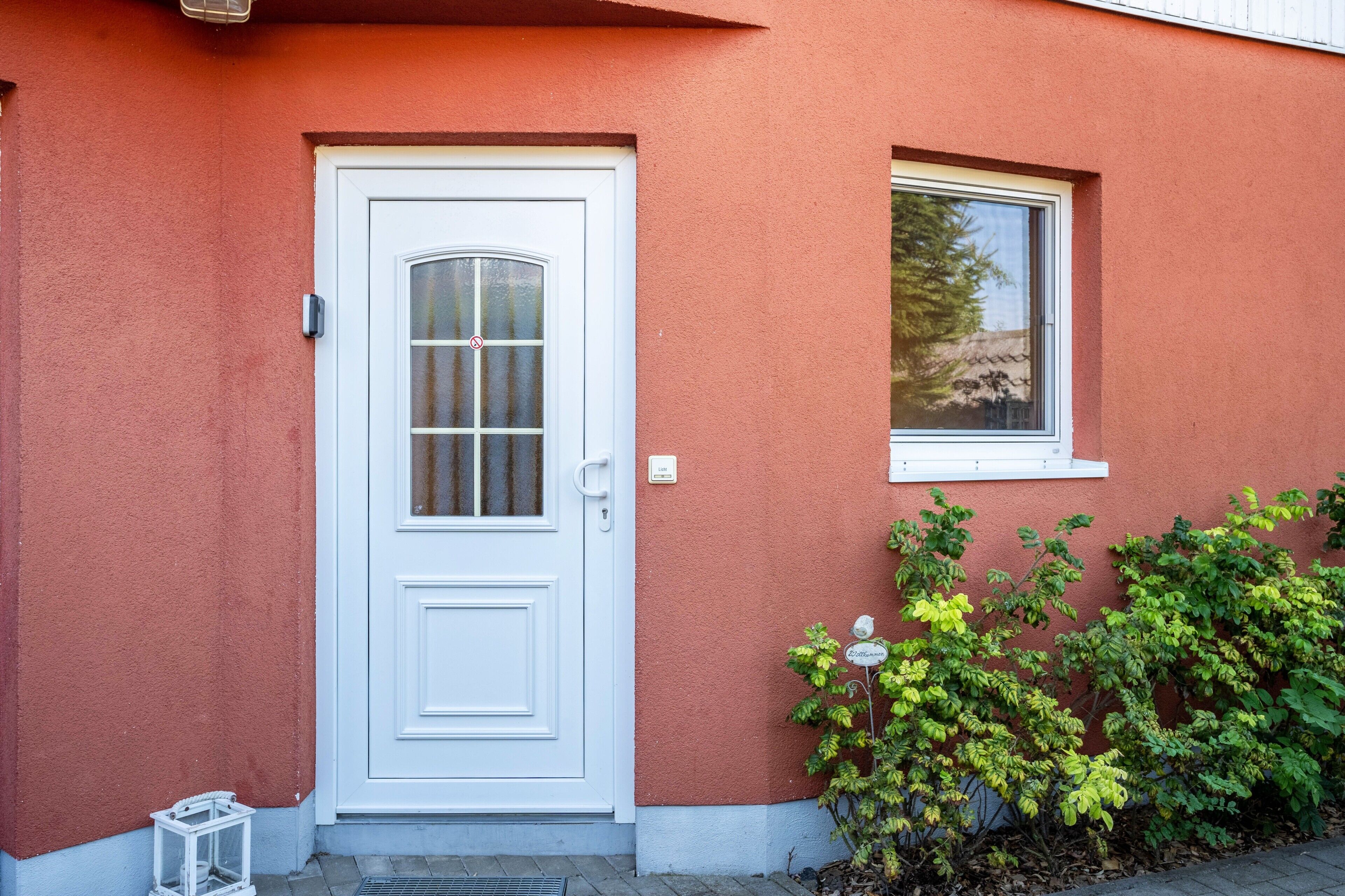 Un bâtiment rouge avec une porte et une fenêtre blanches, une petite lanterne blanche et des feuilles vertes.