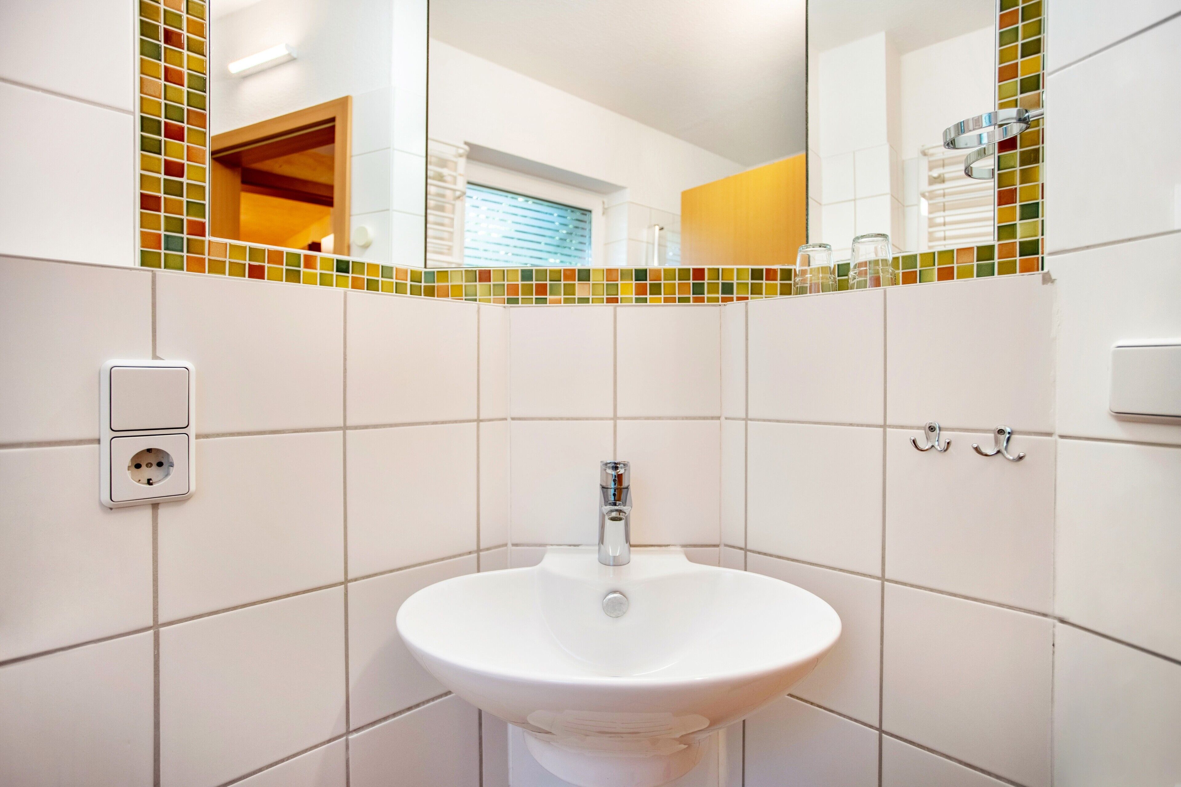 Een badkamer met een witte wastafel, betegelde wanden, een spiegel en een klein stopcontact.