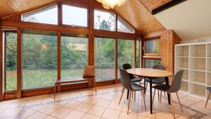 Un comedor con una mesa y sillas de madera, ventanas amplias con persianas y una lámpara de techo.