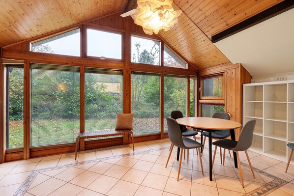 Un espace repas comprenant une table et des chaises en bois, de grandes fenêtres équipées de stores et un luminaire au plafond.