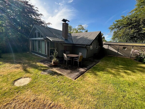 Une maison avec une cheminée, une terrasse avec une table et des chaises, et une cour gazonnée.