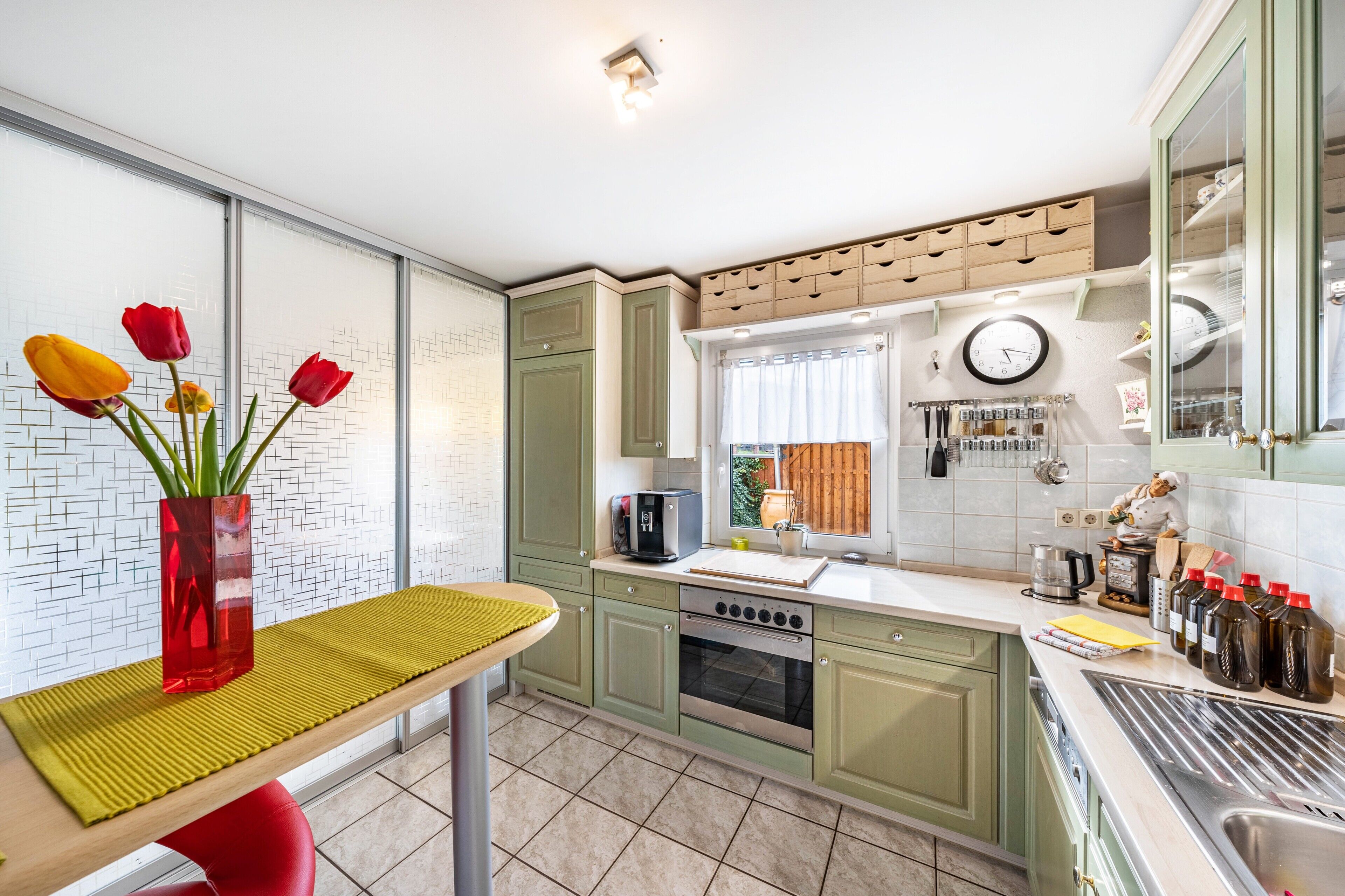 A kitchen with green cabinets, a tiled backsplash, a window with curtains, a clock, and a vase with flowers on the counter.