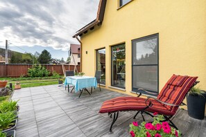 A patio with a table and chairs, a cushioned outdoor seating area, and a yellow building with large windows. - Apartment 'Fewo Energiepunkt Am Planitzwald' with Private Terrace, Private Garden and Wi-Fi (Bennewitz)