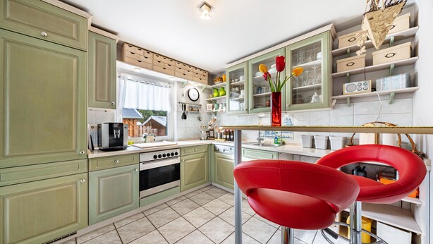 Una cocina con gabinetes verdes, un taburete rojo, un reloj y un florero con flores.