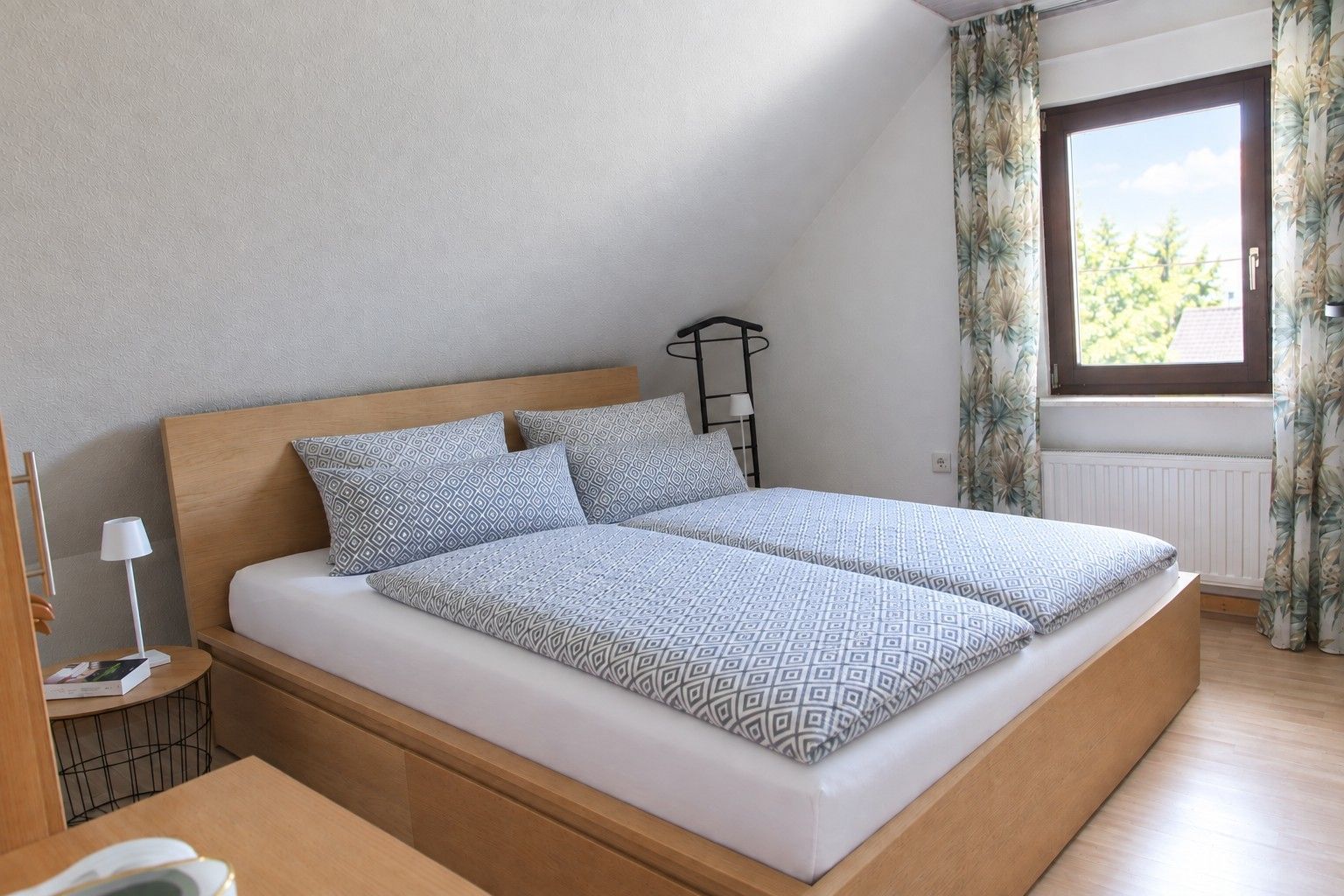 Ein Schlafzimmer mit einem Bett, einem Nachttisch, einer Lampe und einem Fenster mit Vorhängen.