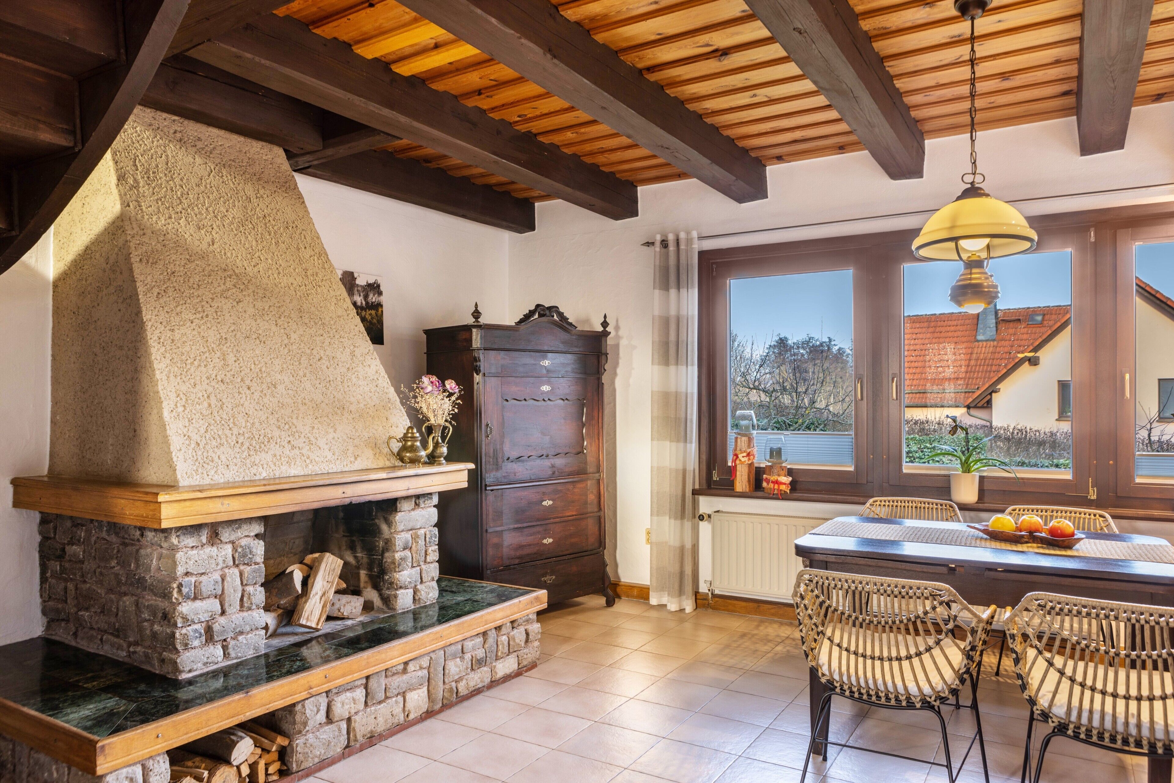 Chimenea de piedra, vigas de techo de madera, una mesa de comedor con sillas y vistas a través de una ventana.