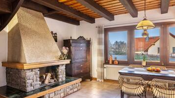 Chimenea de piedra, vigas de techo de madera, una mesa de comedor con sillas y vistas a través de una ventana.