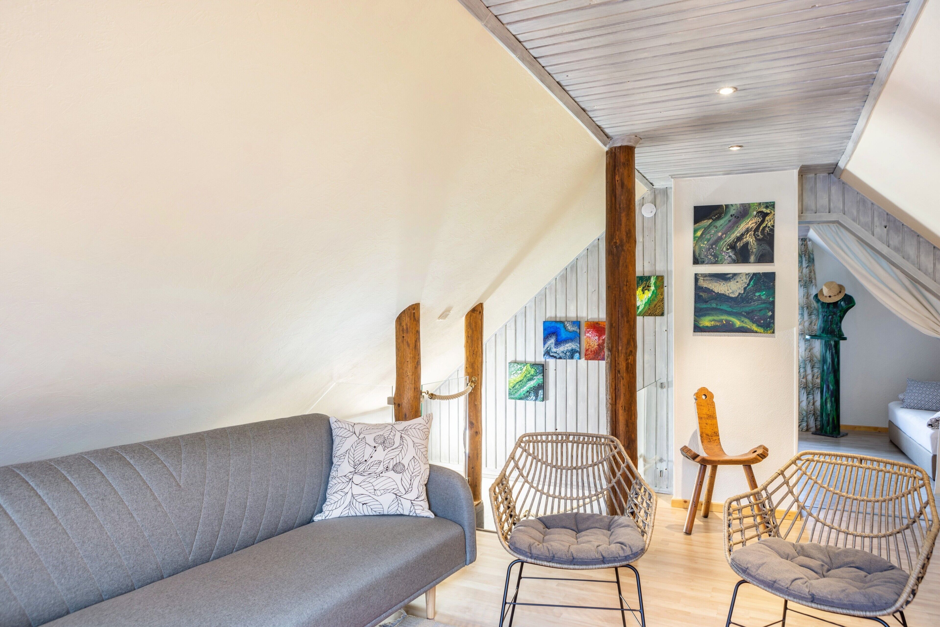 Een loft-achtige kamer met een grijze bank, rieten stoelen, houten balken en kunstwerken aan de muur.
