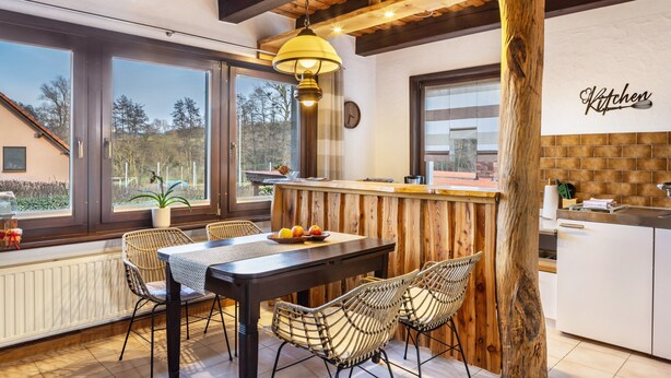 Una cocina moderna con una isla de madera, ventanas amplias, sillas de mimbre y una lámpara colgante.