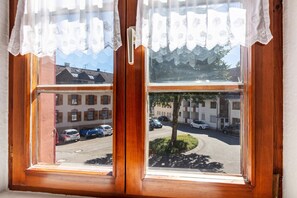 A window with lace curtains, a view of parked cars, and buildings in the background. - Room 'Zimmer 1' with Shared Terrace, Shared Garden and Wi-Fi (Schönau im Schwarzwald)