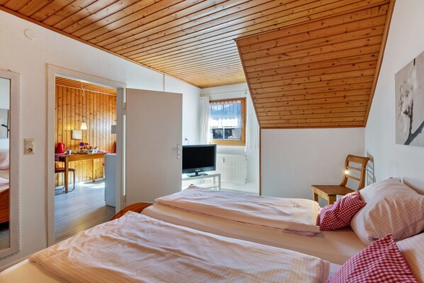 A bedroom with two beds, a wooden ceiling, a TV, and a dining area visible through an open door.